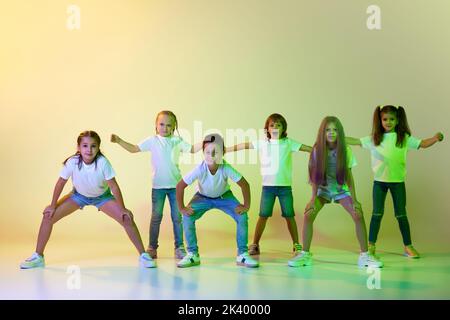 Gruppe von Kindern, aktive Kinder in sportlichen Casual-Stil Kleidung tanzen in Choreographie Klasse isoliert auf grünem Hintergrund in gelbem Neonlicht. Stockfoto