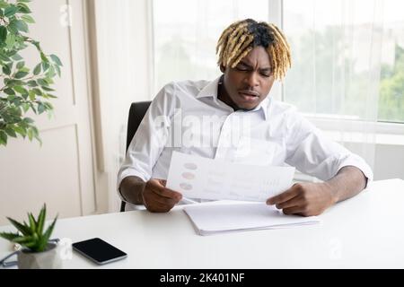 Problem in Betracht ziehen. Besorgter schwarzer Mann. Bericht prüfen. Hübscher, seriöser Typ, der auf dem Schreibtisch sitzt und Informationen in Dokumenten im hellen Innenraum ausgibt. Stockfoto