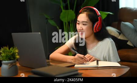 Konzentrierte Frau in Kopfhörer hören Vortrag während des Studiums online auf Laptop und Notizen auf Notebook Stockfoto