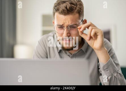 Ein junger bärtiger Mann schaut sich den Laptop genau an, ein fokussierter Mann nutzt den Computer, ein Coder arbeitet zu Hause Stockfoto