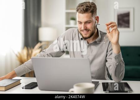 Ein hübscher bärtiger junger Mann, der zu Hause mit Gadgets arbeitet, ein glücklicher Geschäftsmann schaut sich den Laptop an Stockfoto