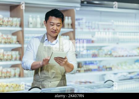 Praktikum bei der Arbeit. Ein junger Mann, ein asiatischer Mann, ein Assistent arbeitet in einem Supermarkt. Auf einer Schürze in der Milchabteilung in der Nähe der Kühlschränke stehend, das Produkt in einem Notebook aufschreiben. Stockfoto