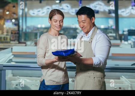 Berater in einem Supermarkt. Ein freundlicher asiatischer Mann in einer Schürze bietet einer jungen schönen Frau an, eine blaue Packung gefrorener Lebensmittel im Laden zu kaufen. Stockfoto