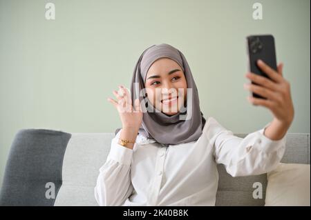 Die schöne, tausendjährige asiatische muslimische Frau mit Hijab-Video ruft ihre Freunde an oder nimmt Videos für ihre sozialen Medien auf ihrem Smartphone auf, während sie darin sitzt Stockfoto