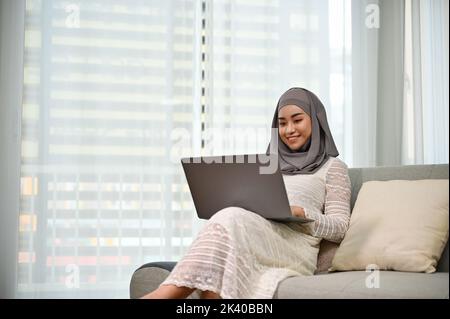 Schöne und elegante tausendjährige asiatische muslimische Frau oder Geschäftsfrau mit Hijab mit Laptop-Computer, um ihre Aufgaben zu verwalten, während sie sich auf ihrem Comfor entspannen Stockfoto