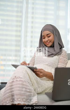 Porträt, schöne tausendjährige asiatische muslimische Frau trägt Hijab mit digitalen Tablet, während sie sich in ihrem Wohnzimmer entspannen. Stockfoto