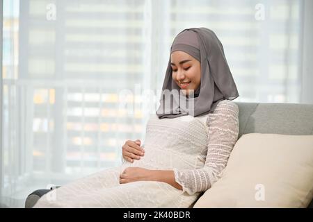 Schöne und glückliche, tausendjährige asiatische muslimische Schwangerin, die ihren Bauch berührt und auf ihrer bequemen Couch im Wohnzimmer sitzt. Mutterschaft, Pregna Stockfoto