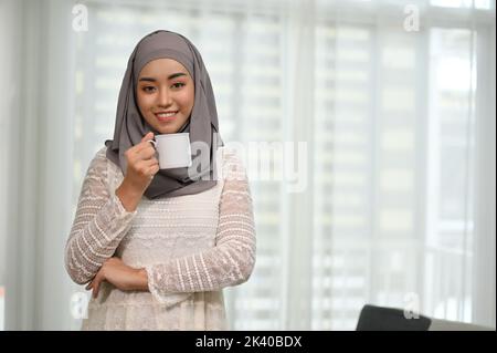 Attraktive und elegante, tausendjährige asiatische muslimische Frau, die in ihrem Wohnzimmer in einem Hijab mit einer Tasse Kaffee steht. Stockfoto