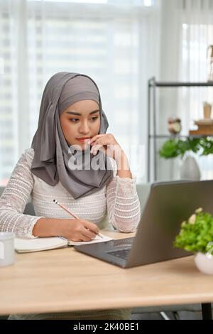 Porträt, nachdenklich junge asiatische muslimische weibliche College-Studentin nachdenklich über ihre Schulaufgabe, Hausaufgaben auf ihrem Laptop-Computer in h Stockfoto