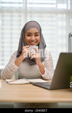 Porträt, schöne und attraktive tausendjährige asiatische muslimische Frau trägt Hijab genießen ihren Kaffee, während sie an ihrem Schreibtisch arbeiten. Stockfoto