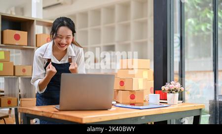 Erfolgreiche und fröhliche junge asiatische weibliche kleine Online-Geschäftsbesitzerin glücklich mit ihren Mid-month-Bestellungen, Überprüfung der Verkäufe auf ihrem Laptop-Computer, feiern Stockfoto