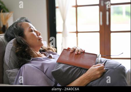 Friedliche und entspannte, gealterte asiatische Frau schläft ein, während sie ein Buch auf ihrem bequemen Sofa in ihrem Wohnzimmer liest. Lifestyle-Konzept der Rentnerin Stockfoto
