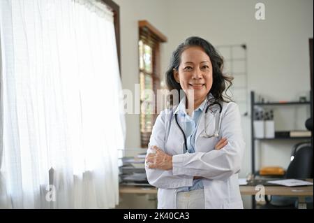 Intelligente und professionelle asiatische Senior-Ärztin in weißer Uniform, Arme gekreuzt, steht in der Arztpraxis. Stockfoto