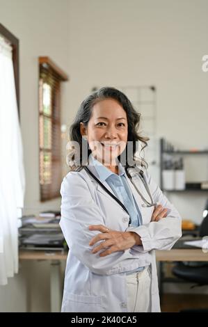 Portrait, Smart und professionelle asiatische Senior-Ärztin in weißen Gewand Uniform, Arme gekreuzt, steht in der Arztpraxis. Stockfoto