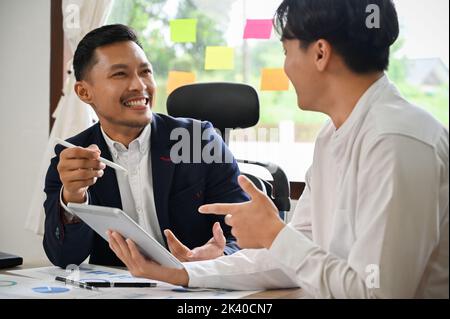 Zwei asiatische männliche Geschäftskollegen oder Geschäftsleute unterhalten sich gern, teilen Geschäftsideen, während sie an ihrem Projekt zusammenarbeiten. Stockfoto