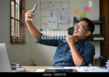 Fröhlicher und fröhlicher asiatischer Geschäftsmann oder Büroangestellter, der Selfie macht oder gerne mit seinem Freund Duri über einen Videoanruf auf seinem Smartphone spricht Stockfoto