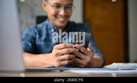 Glücklicher asiatischer Geschäftsmann oder Büroangestellter mit Smartphone, plaudert gern mit seinen Freunden oder schaut sich während des Arbeitsaufenthalten etwas Lustiges im Internet an Stockfoto