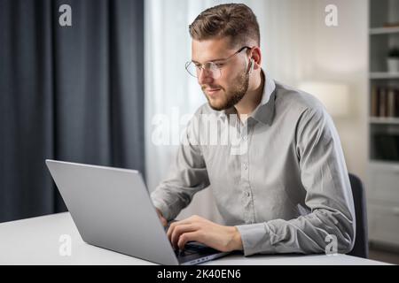 Schöner Mann in grauem Hemd, der von zu Hause aus am Laptop codiert und fernab arbeitet, um Code zu schreiben Stockfoto