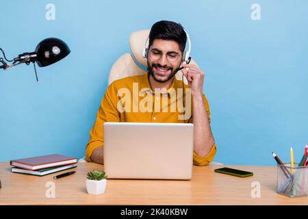 Foto von positiven professionellen Betreiber Mann sitzend Stuhl Hand-Touch-Kopfhörer sprechen Client isoliert auf blauem Hintergrund Stockfoto