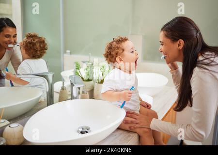 Mutter lehrt Baby, seine Zähne zu putzen, auf der Badezimmertheke zu Hause und ein sauberes Lächeln auf ihrem Gesicht. Gesunde Mundhygiene für Kinder bedeutet, Kinder zu verwenden Stockfoto