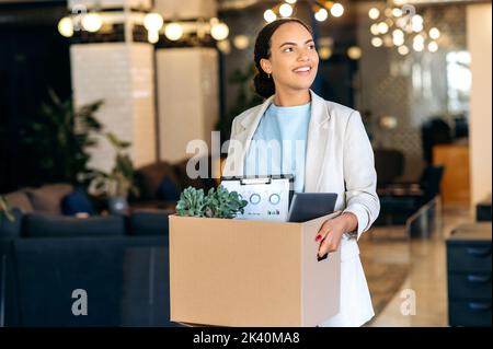 Neuer Job. Eine begeisterte, erfolgreiche, motivierte Mixed-Race-Frau, die in ein neues Arbeitsbüro geht, einen Karton mit Arbeitsattributen in den Händen hält, erstaunt den Arbeitsplatz inspiziert und glücklich lächelt Stockfoto