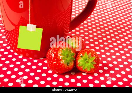 Rote Tasse mit Teebeutel und Erdbeeren auf rot-weißen Tupfen Hintergrund wie fröhliche Concet von Früchtetee Stockfoto