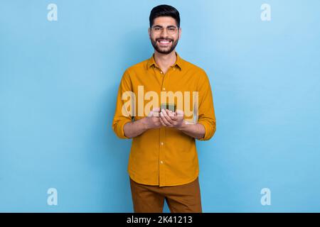 Foto von smart clever jungen positiven Mann gekleidet gelben Hemd in Brille halten in den Händen Smartphone isoliert auf blauem Hintergrund Stockfoto