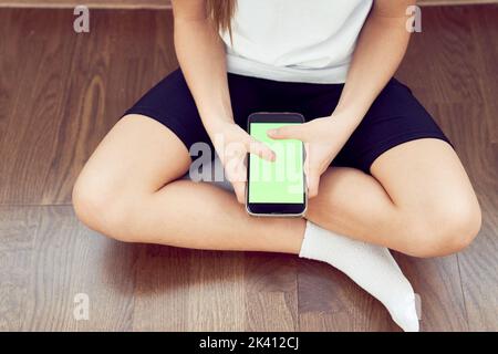 Hände von Kindern mit einem grünen Bildschirm. Ein kleines Mädchen sitzt auf dem Boden und hält ein Smartphone mit einer Chromakey in den Händen. Blick aus einem großen Winkel. Hochwertige Fotos Stockfoto