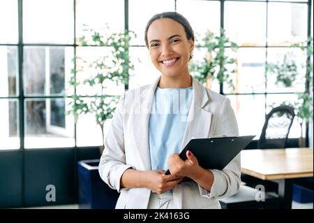 Porträt eines erfolgreichen zufriedenen schönen Mixed-Race-Geschäft bereit, Einstellung Manager, Broker, hält einen Ordner mit Dokumenten, im Büro stehen, Blick auf die Kamera, lächelnd freundlich Stockfoto