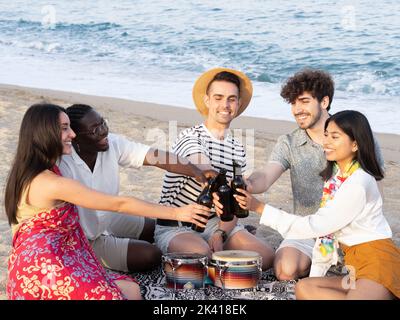 Gruppe von fröhlichen, multiethnischen jungen Menschen am Strand, die mit Bier toasten. Beste Freunde, die Spaß haben. Hochwertige Fotos Stockfoto