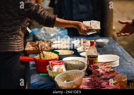 Nahaufnahme einer Frau, die ihre Hand mit vietnamesischem Bargeld ausstreckt, um eine Schüssel Nudeln für das Mittagessen in der Markthalle von Meo Vac zu kaufen. Stockfoto