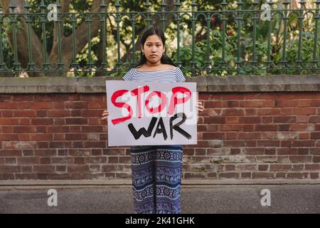 Ein junges Mädchen im Teenageralter, das die Kamera anschaut, während es ein Anti-Kriegs-Plakat hält. Junge Friedensaktivistin, die gegen Krieg und Gewalt protestiert. Stockfoto