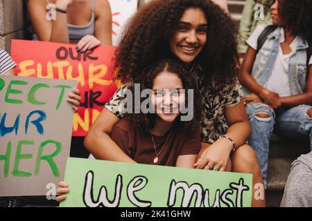 Fröhliche Mädchen im Teenageralter lächeln an der Kamera, während sie mit einer Menge von Klimaaktivisten sitzen. Gruppe von multikulturellen Demonstranten, die für das Klima streiken j Stockfoto