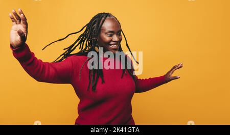 Fröhliche reife Frau, die tanzt und Spaß hat, während sie vor einem gelben Hintergrund steht. Fröhliche schwarze Frau mit Dreadlocks lacht fröhlich in einem Gestüt Stockfoto