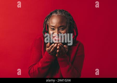 Reife Frau lächelt und weht Luft Konfetti, während sie vor einem roten Hintergrund steht. Glückliche Frau mittleren Alters mit Dreadlocks, die sich in einem Studio amüsieren. Stockfoto