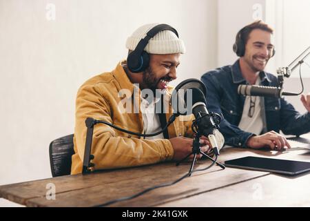 Zwei College-Podcasters lachen und haben eine gute Zeit in einem Studio. Zwei glückliche junge Männer, die gemeinsam eine Live-Audioübertragung moderiert haben. Zwei männliche Inhaltsersteller r Stockfoto