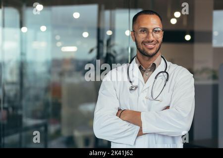 Porträt eines jungen glücklichen und lächelnden Arztes, eines Mannes im medizinischen Mantel und Stethoskop, der lächelt und die Kamera anschaut, eines arabischen Arztes mit gekreuzten Armen, der im modernen Klinikbüro arbeitet. Stockfoto