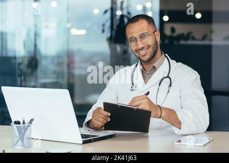 Porträt eines erfolgreichen männlichen Arztes, hispanisch lächelnd und mit Blick auf die Kamera, Arzt im medizinischen Gewand, der im modernen Klinikbüro arbeitet und Laptop für die Arbeit verwendet Stockfoto
