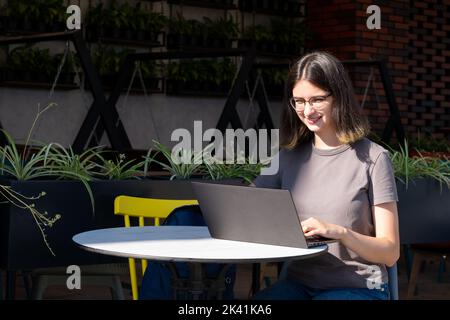 Freiberufliches Konzept. Eine schöne Brünette Student Mädchen mit Brille verwendet einen Laptop in einem Straßencafe, schöne Schatten. Schöne Mädchen Student mit a la Stockfoto
