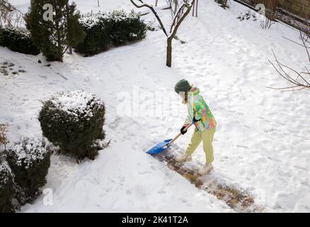 Eine nicht erkennbare junge Frau in strahlend warmer Sportkleidung räumt nach einem schweren Schneesturm mit einer großen Seitenschaufel den Weg im Hof vom Schnee frei. Winter s Stockfoto