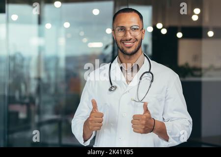 Erfolgreicher afroamerikanischer Arzt, der die Kamera ansah und lächelte und die Daumen hoch hielt, ein Mann in einem medizinischen Mantel und einer Brille mit Stethoskop, der im modernen Klinikbüro arbeitete. Stockfoto