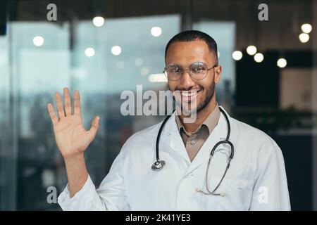 Junger afroamerikanischer Arzt, der eine Brille trägt und einen medizinischen Mantel trägt, der glücklich und lächelnd grüßt. Freundliche Geste, freundlicher Arzt, der in der Klinik arbeitet. Stockfoto