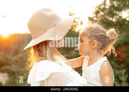 Nicht erkennbare lächelnde Frau in Hut umarmt kleine blonde Mädchen, Tochter auf natürlichen unscharfen Hintergrund. Kindersicherheit Stockfoto