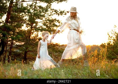 Hübsche Frau, Mutter im Hut und kleine blonde Tochter in hellweißen Kleidern, die gegen den Sonnenuntergang im Wald zusammenlaufen. Stockfoto