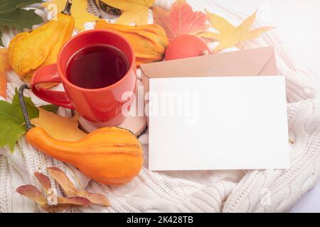 Unbeschriftete Karte mit Umschlag, Tasse Tee, orangefarbenen Kürbissen und Herbstblättern auf weißem Stricktuch. Thanksgiving, Herbstkonzept. Draufsicht, Nachbau. Stockfoto