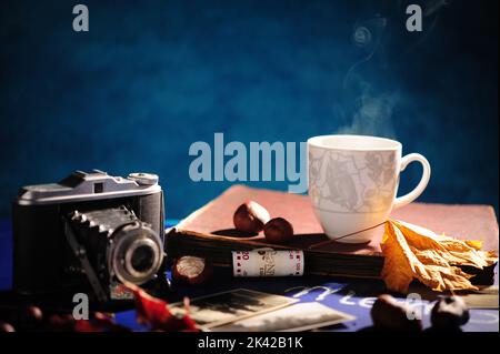 Kaffeedampf aus Tasse, Büchern, Vintage-Kamera und Herbstblättern. Horizontales Foto mit blauem Hintergrund und Platz zum Kopieren nach links. Stockfoto