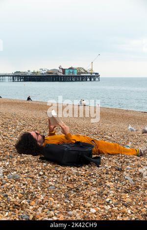 Junger Mann, der am Strand liegt und einen Taschenbuchroman liest, entspannt sich auf Sand und Kieselsteinen mit dem Meer und dem Pier des Brighton Palace in der Ferne. VEREINIGTES KÖNIGREICH. (131) Stockfoto