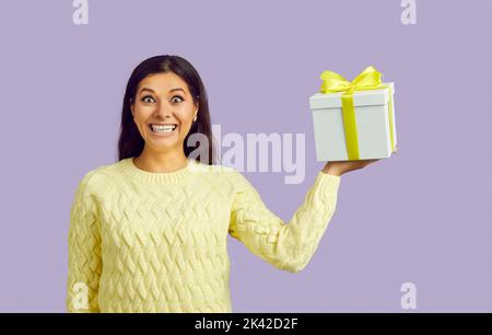 Aufgeregt lustige junge Frau mit fröhlichem verrückten Lächeln hält Geschenkbox auf pastelllila Hintergrund. Stockfoto