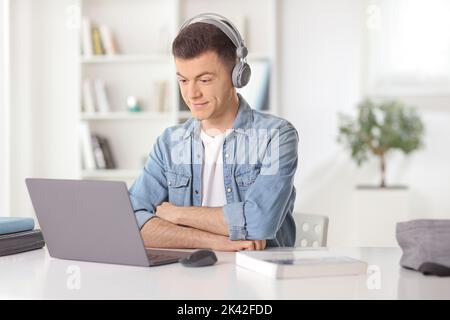 Junger Mann sitzt zu Hause vor einem Laptop und hört sich eine Anleitung mit Kopfhörern an Stockfoto