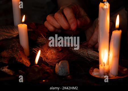 Voodoo Puppe in den Händen einer alten Hexe mit einer Nadel im Herzen im Dunkeln vor dem Hintergrund von Kerzen, Flüchen und schwarzer Magie Stockfoto